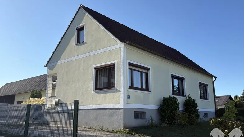 Einfamilienhaus mit heller Fassade, Satteldach und braunen Fensterläden unter blauem Himmel.