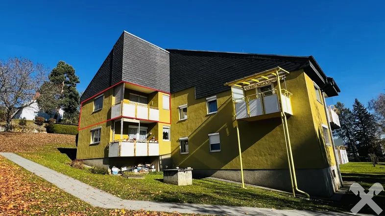 Mehrfamilienhaus mit gelber Fassade, Balkonen und Mansarddach in grüner Umgebung.