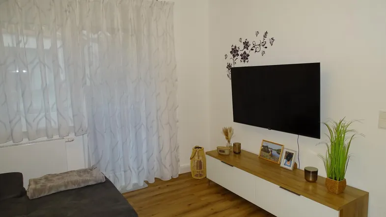 Gemütliches Wohnzimmer mit Fernseher, modernem Sideboard und großen Fenstern.