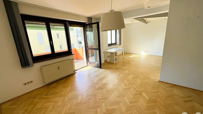 Heller Wohnbereich mit Fischgrätparkett und Zugang zu einem sonnigen Balkon, bietet viel Tageslicht.
