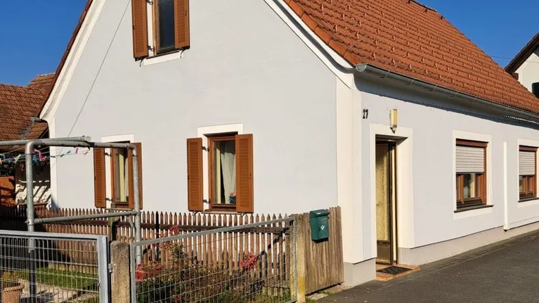 Traditionelles Einfamilienhaus mit hellgrauer Fassade, braunen Fensterläden und Ziegeldach.