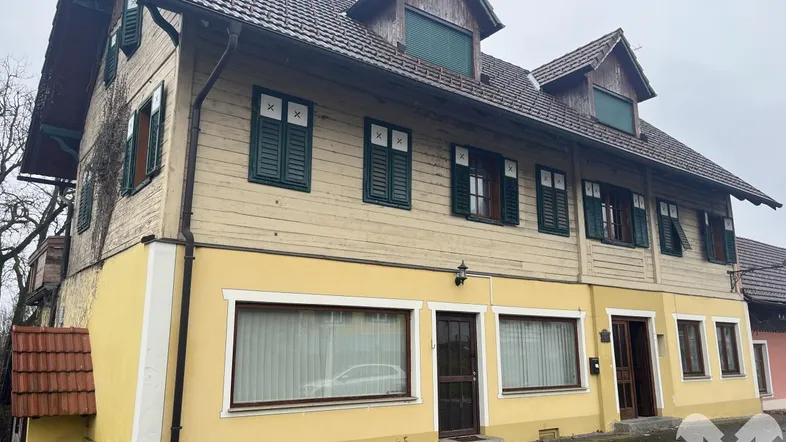 Ansicht der Gebäudefassade mit gelbem Putz, Holzverkleidung und traditionellen Fensterläden an einem bewölkten Tag.