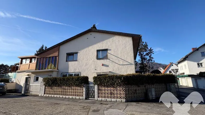 Vorderansicht des Gebäudes mit weißer Fassade, Holzelementen und gepflegter Hecke.