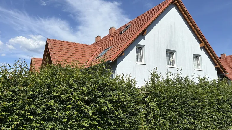 Reihenhausfassade mit hellblauer Farbe und rotem Ziegeldach, umgeben von einer grünen Hecke unter blauem Himmel.