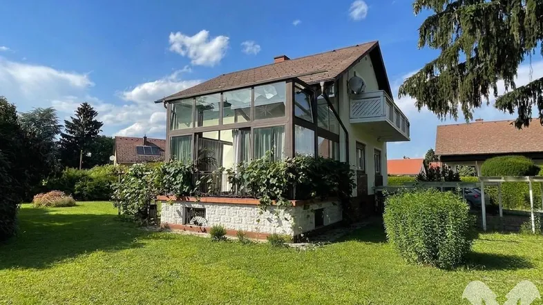 Freistehendes Haus mit großem verglastem Wintergarten und gepflegtem Garten unter blauem Himmel.