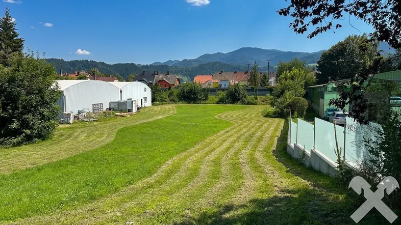 Große grüne Wiese mit gemähten Bahnen, umgeben von Gebäuden und Bergen im Hintergrund unter blauem Himmel.