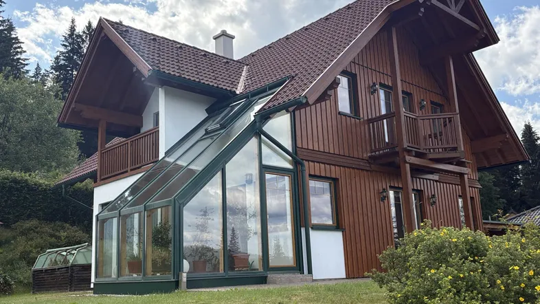 Charmantes Haus mit Holzfassade, Wintergarten und Satteldach, eingebettet in grüne Landschaft.