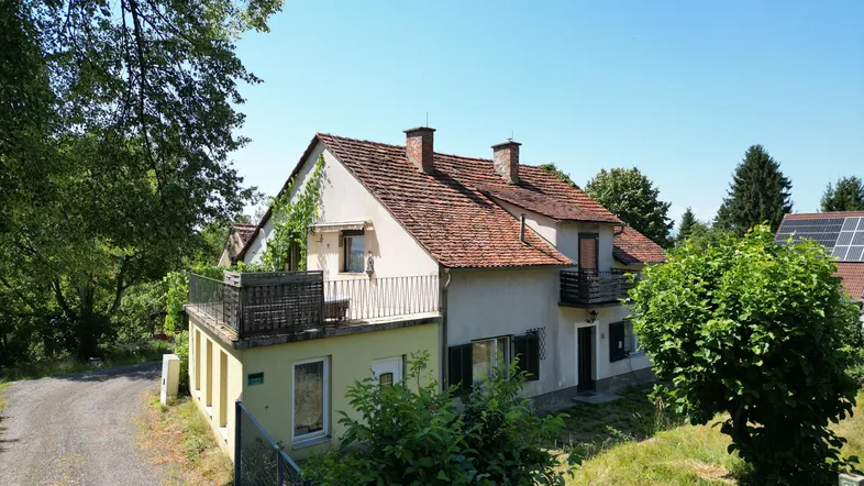 Hausfassade mit Balkon und üppiger Vegetation in ländlicher Umgebung unter blauem Himmel.