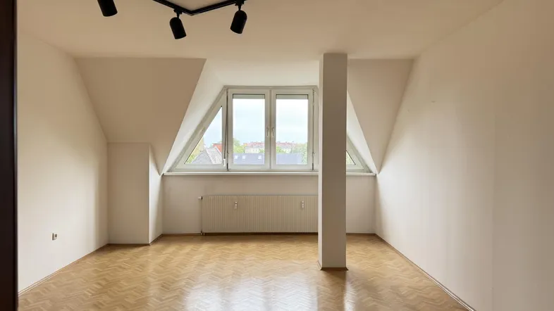Helles Zimmer im Dachgeschoss mit Parkettboden, schrägen Wänden und einem großen Fenster mit Stadtblick.