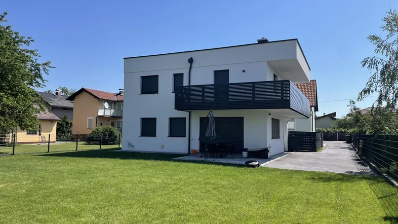 Moderne weiße Hausfassade mit großem Rasenbereich und gepflasterter Terrasse im Freien.