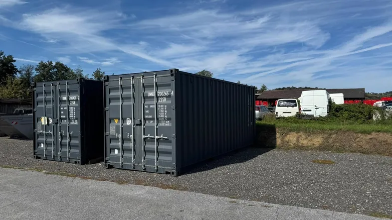 Zwei dunkelgraue Schiffscontainer stehen auf einem Schotterplatz unter blauem Himmel.