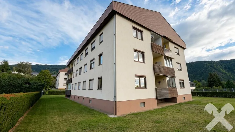 Mehrfamilienhaus mit braunem Dach und heller Fassade, umgeben von Grünflächen und Bergen.