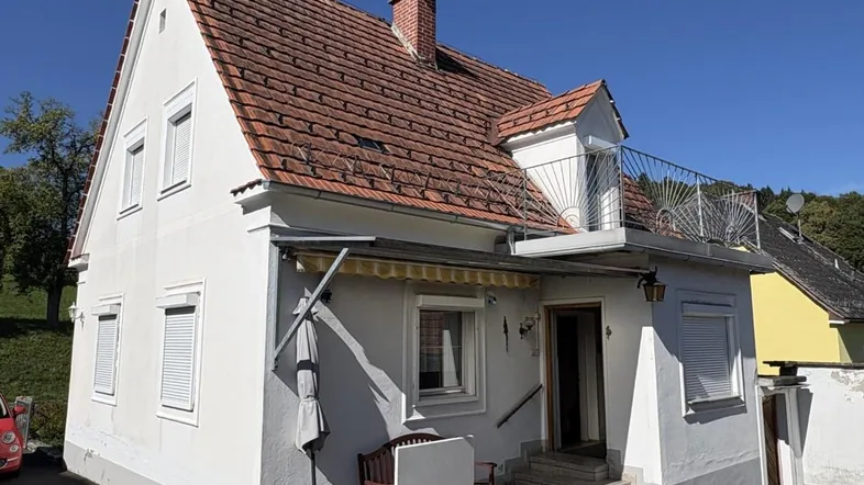 Vorderansicht des Hauses mit rotem Ziegeldach, kleinem Balkon und einer gepflasterten Terrasse mit Sitzgelegenheit.