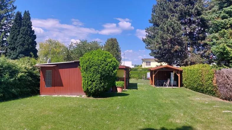 Gepflegter Garten mit Rasenfläche, Gartenhaus und einem überdachten Sitzbereich unter blauem Himmel.