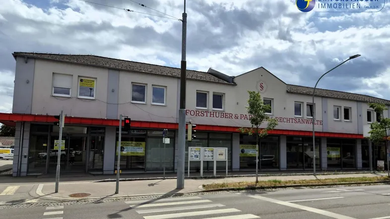 Gebäudeansicht von der Straße mit großen Schaufenstern und einem Zebrastreifen davor unter bewölktem Himmel.