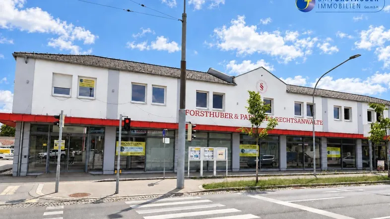 Gebäudeansicht eines gepflegten Geschäftshauses mit weißer Fassade, roten Markisen und großen Schaufenstern an einer Straße.