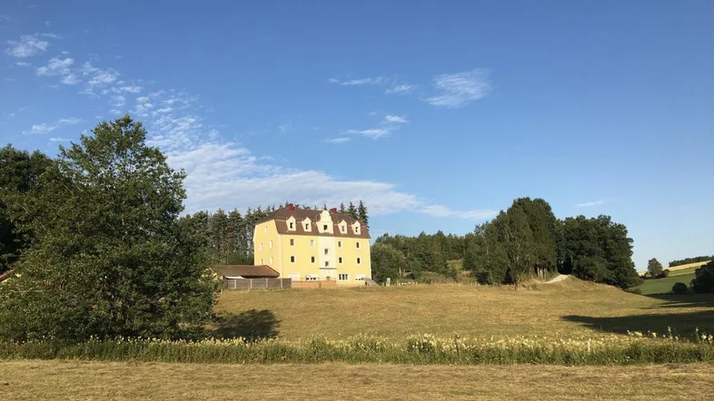 Gelbes Gebäude mit Mansarddach inmitten einer weiten grünen Landschaft unter blauem Himmel.
