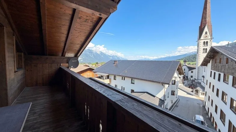 Geräumiger Holzbalkon mit Blick auf das Dorf, die Berge und einen Kirchturm unter blauem Himmel.