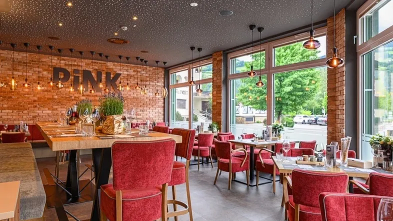 Geräumiger Restaurantbereich mit freiliegenden Ziegelwänden, großen Fenstern und roten Stühlen, die eine einladende Atmosphäre schaffen.
