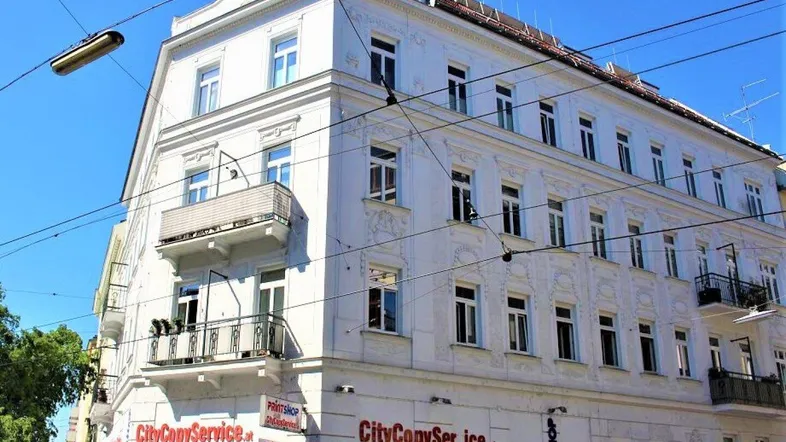 Historisches Gebäude mit weißer Fassade, dekorativen Elementen und mehreren Balkonen in städtischer Umgebung.
