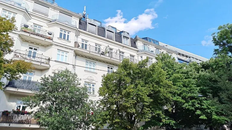 Klassisches Mehrfamilienhaus mit Balkonen und grünen Bäumen unter blauem Himmel.