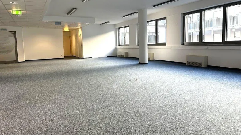 Geräumiger, leerer Büroraum mit Teppichboden und großen Fenstern, ideal für flexible Arbeitsplatzgestaltung.