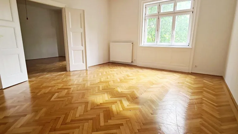 Helles Zimmer mit Fischgrätparkettboden, weißen Wänden und großem Fenster mit Blick ins Grüne.