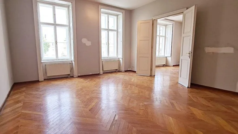 Geräumiges Zimmer mit Fischgrätparkett und hohen Fenstern, das in einen weiteren Raum führt.