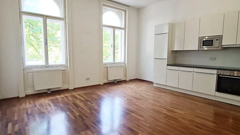 Heller Wohnbereich mit integrierter weißer Küche, Parkettboden und hohen Bogenfenstern.