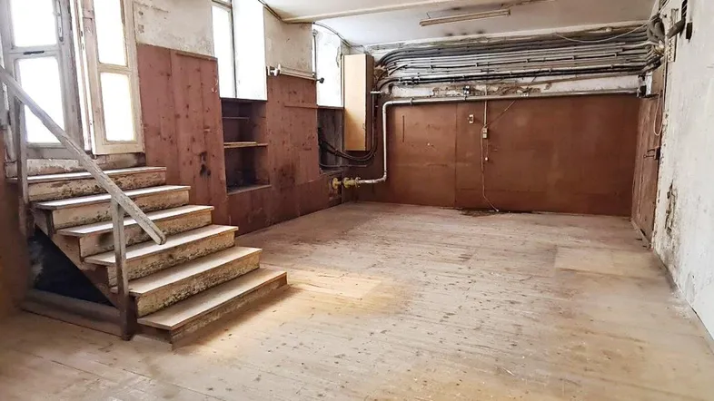 Großer, leerer Raum mit Holzdielenboden, sichtbaren Rohren und einer Holztreppe, die nach oben führt.