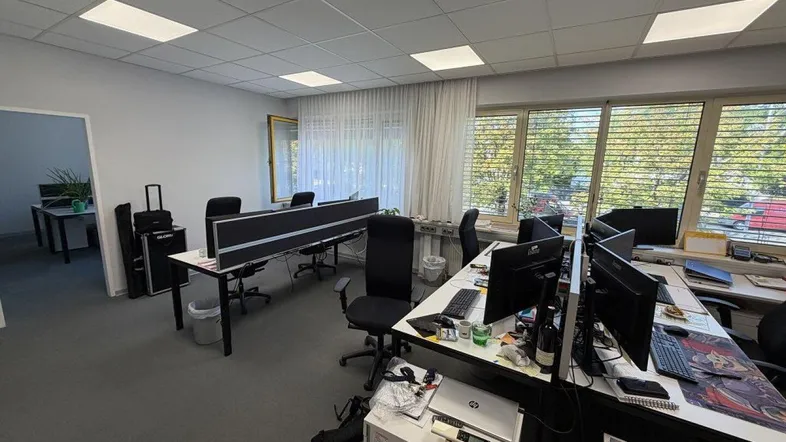 Großraumbüro mit mehreren Arbeitsplätzen, Teppichboden und großen Fenstern, die viel Tageslicht hereinlassen.