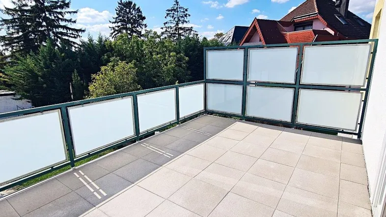 Geräumige Terrasse mit Fliesenboden und moderner Glasbrüstung unter blauem Himmel.