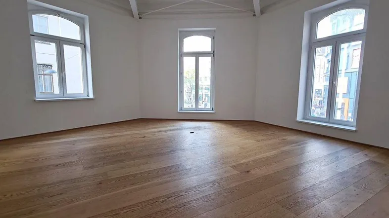 Geräumiger, heller Raum mit Holzboden, hohen Decken und drei großen Bogenfenstern, die viel Licht hereinlassen.
