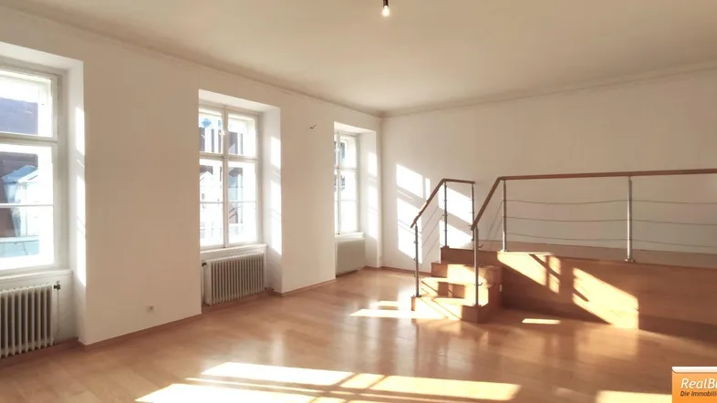 Helles Wohnzimmer mit Holzparkettboden, großen Fenstern und einer Treppe zu einem erhöhten Bereich.