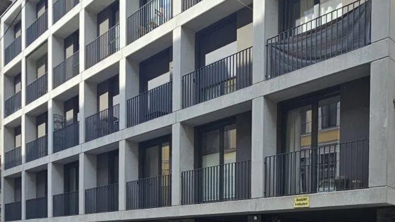 Moderne Wohnhausfassade mit klaren Linien und Balkonen, davor parkende Fahrzeuge auf der Straße.