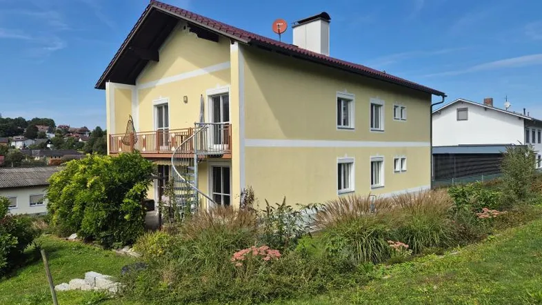 Gelbes Einfamilienhaus mit Balkon, Wendeltreppe und gepflegtem Gartenbereich unter blauem Himmel.