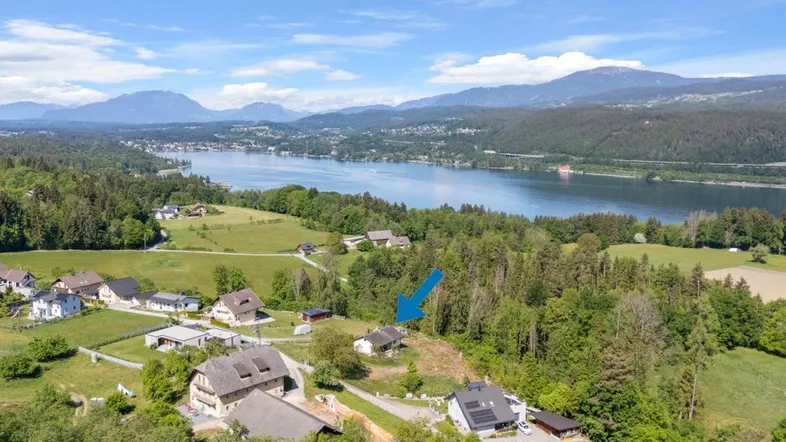 Luftaufnahme eines Hauses mit Blick auf einen See und die umliegende Berglandschaft, markiert mit einem blauen Pfeil.