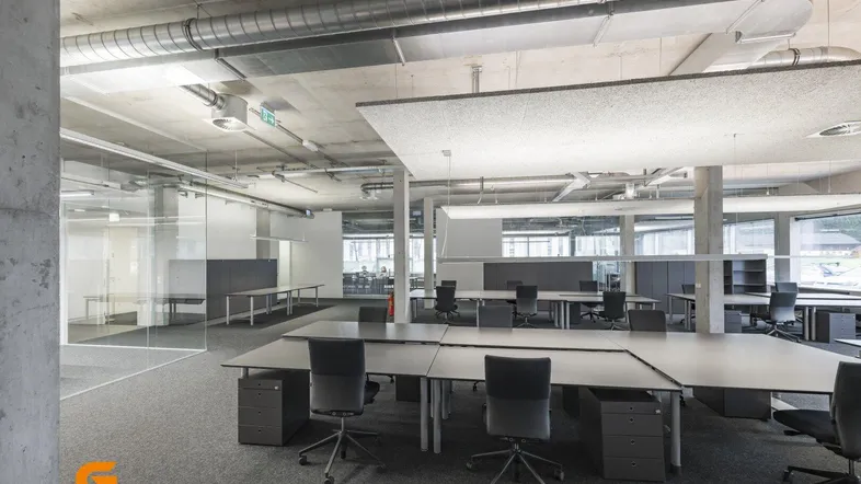 Modernes Großraumbüro mit Sichtbetondecke, freiliegenden Lüftungsrohren und Glasabtrennungen für Arbeitsbereiche.