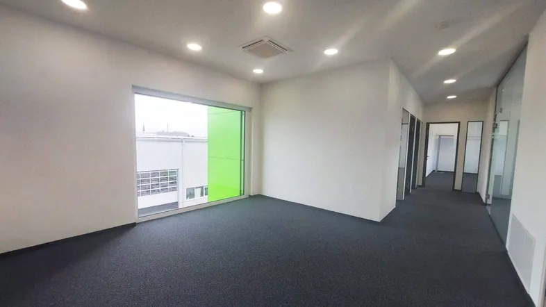 Geräumiges, leeres Büro mit großen Fenstern, dunklem Teppichboden und moderner Beleuchtung.