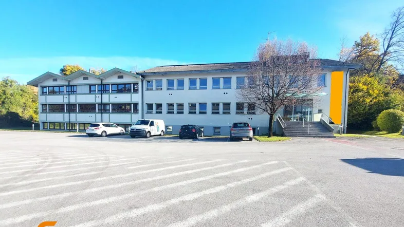 Gebäudeansicht eines Gewerbeobjekts mit großem Parkplatz unter blauem Himmel und umgebender Natur.