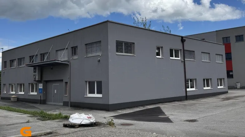 Modernes graues Gebäude mit vielen Fenstern und einem gepflasterten Vorplatz unter bewölktem Himmel.