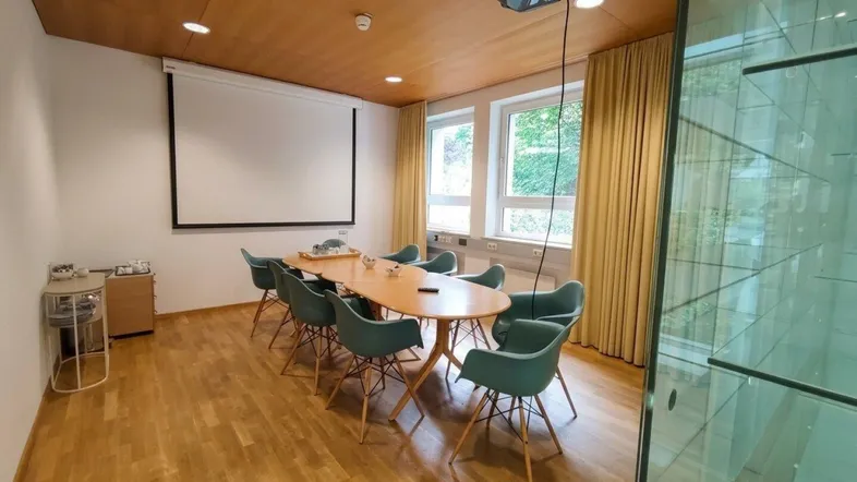 Moderner Konferenzraum mit Holzboden, Projektor, Leinwand und Blick ins Grüne durch große Fenster.
