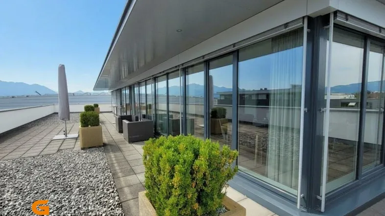 Große Dachterrasse mit moderner Architektur, raumhohen Fenstern und Panoramablick auf die Berge.