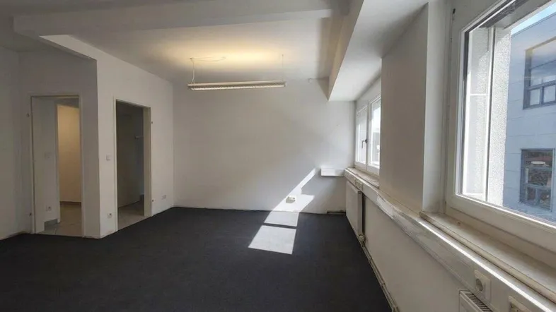 Heller Büroraum mit dunklem Teppichboden und großen Fenstern, die viel Tageslicht hereinlassen.