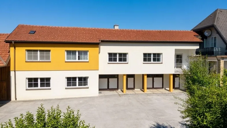 Gebäudeansicht mit gelber und weißer Fassade, rotem Ziegeldach und großem Vorplatz.