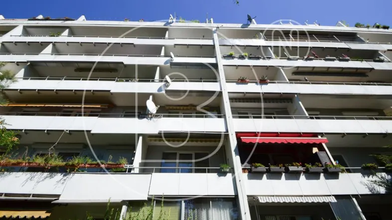 Fassade eines Apartmentgebäudes mit mehreren Balkonen, Markisen und Pflanzenkästen unter blauem Himmel.