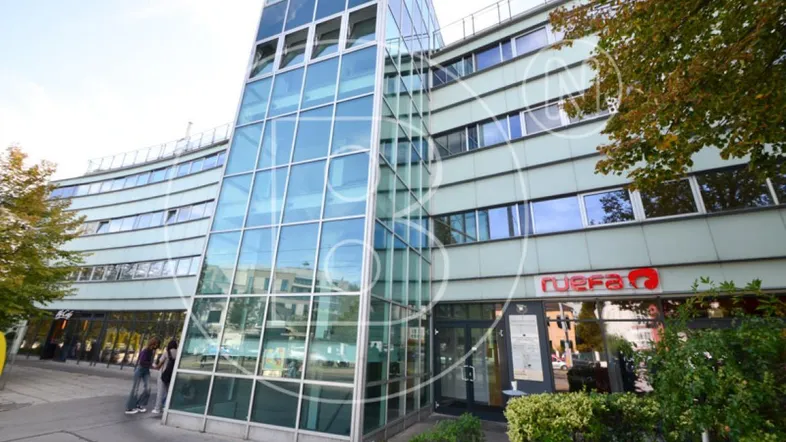 Modernes Bürogebäude mit großer Glasfassade und Eingangsbereich, umgeben von Bäumen.