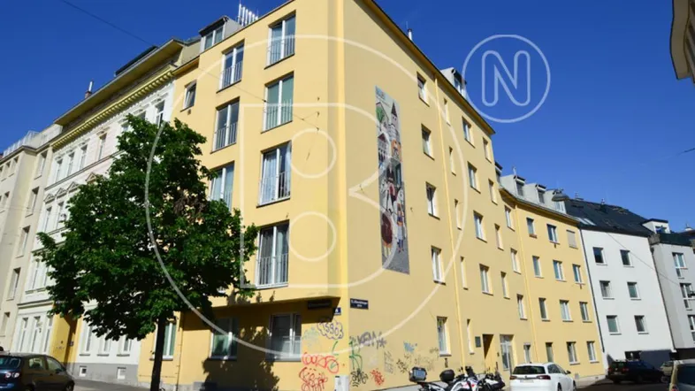 Gelbes Mehrfamilienhaus mit mehreren Stockwerken und Fenstern an einer sonnigen Straße.