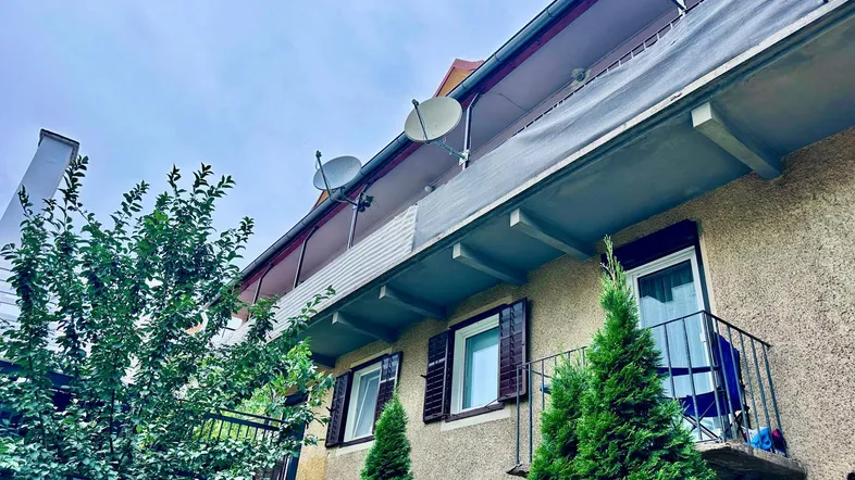 Gebäudefassade mit mehreren Balkonen, Satellitenschüsseln und grünen Bäumen unter bewölktem Himmel.