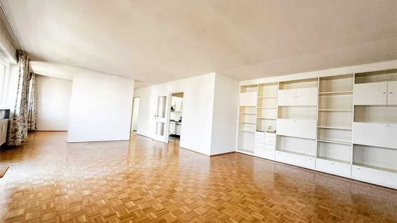 Geräumiges Wohnzimmer mit Parkettboden und integrierten weißen Regalen, offen zur Küche.